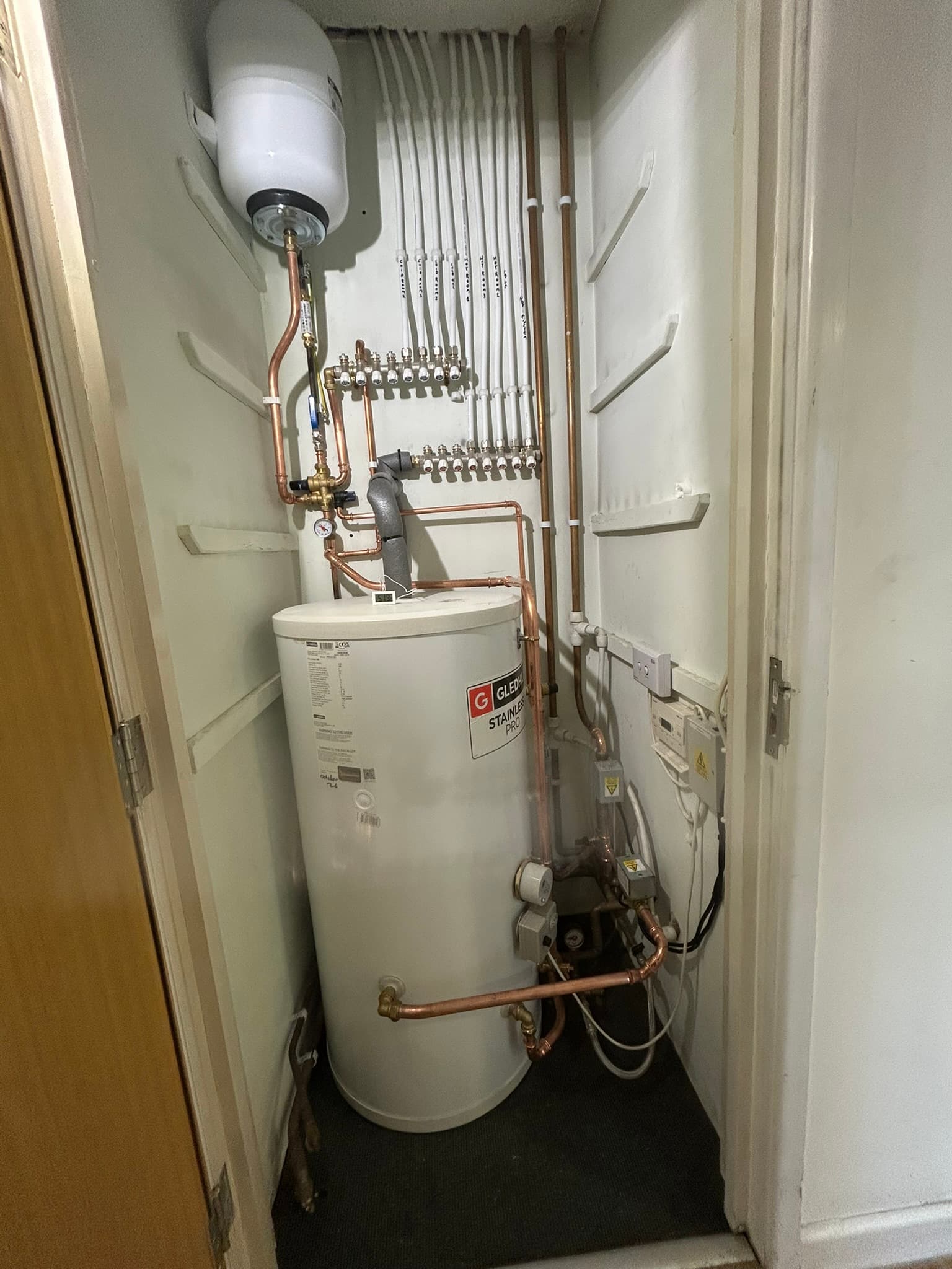 Cupboard in a doctors' surgery with a new white unvented hot water cylinder, an expansion vessel and two manifolds feeding white pipes, each labelled with the room it serves