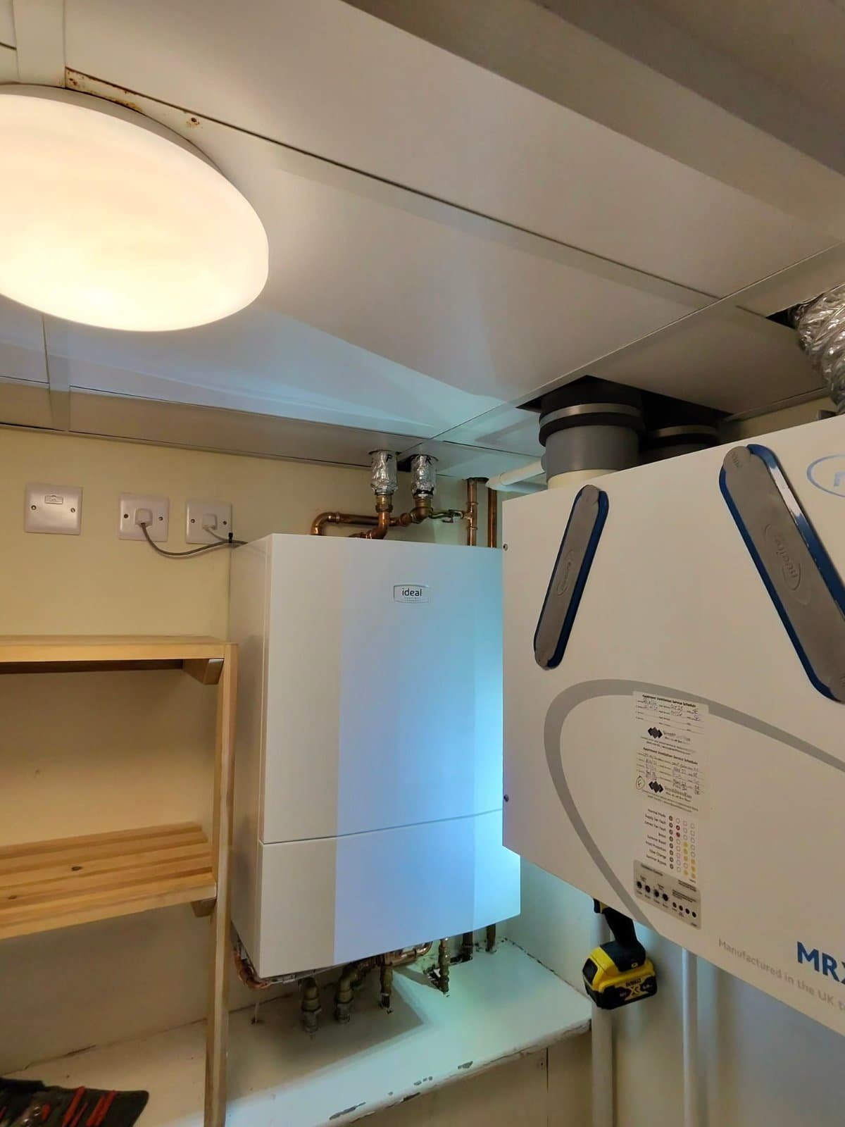 New white Ideal heat interface unit fitted in a utility cupboard, with insulated pipework above it, a wooden shelf unit to the left and a ventilation unit to the right